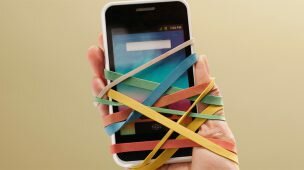 Imagem de mão amarrada a um smartphone com ajuda de elásticos coloridos para ilustrar conteúdo sobre dependência tecnológica.